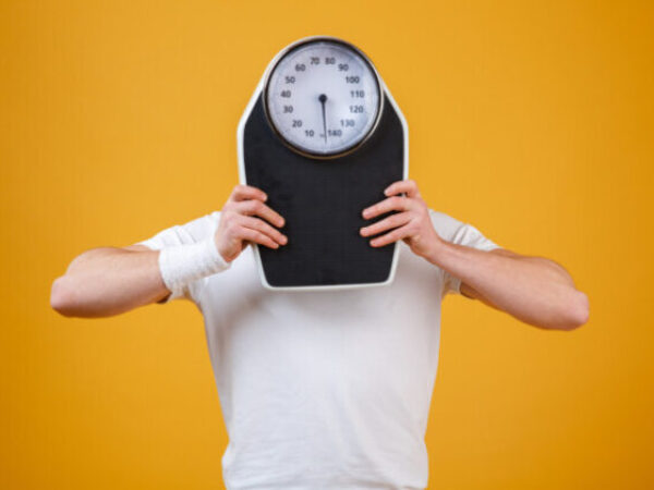 Young sports man hiding face behind weight scales isolated on orange background